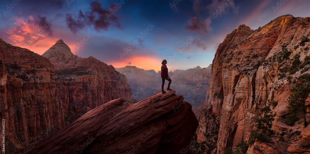 Adventurous Woman at the edge of a cliff is looking at a beautiful landscape view in the Canyon during a vibrant sunset. Taken in Zion National Park, Utah, United States. Sky Composite Panorama