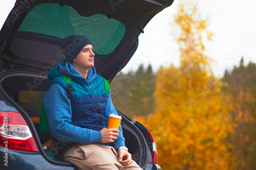 Close-up, a man sits on the trunk of his car in the woods. Male camping in the forest, autumn landscape, A glass of coffee to go