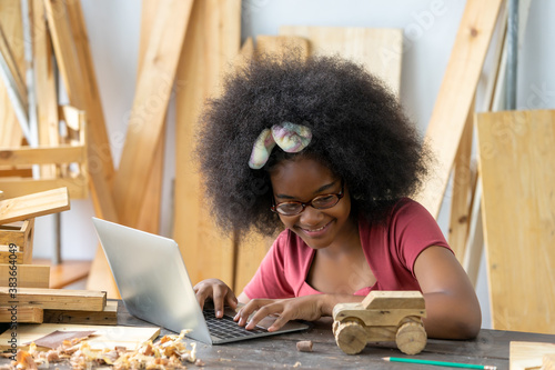 A little inventor working as a woodworker uses the computer to craft woodworking at a workshop. African American girl practice to be a carpenter in a carpentry shop. DIY crafts and Hobbies concepts photo