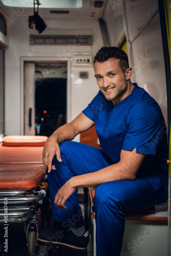 A portrait of a smiling paramedic in a uniform sitting in the back of an ambulance with a stretcher.
