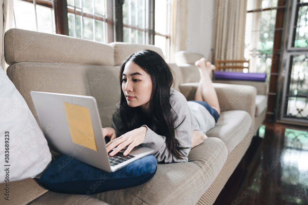Naklejka premium oung adult asian woman laying on sofa with laptop computer for work and online media at home