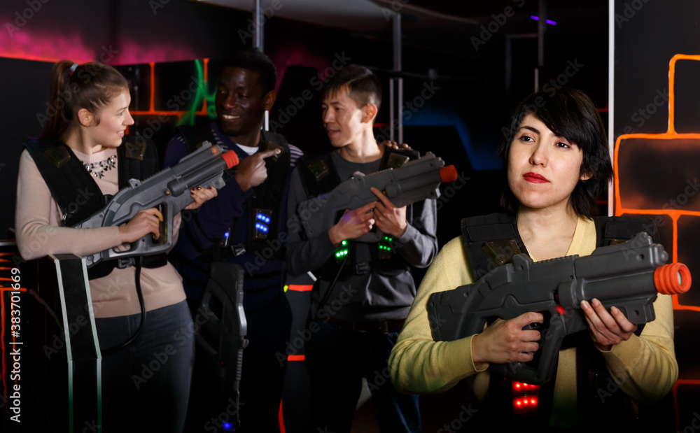 Smiling Aztec woman wearing special vest and holding laser gun ready for lasertag game with friends indoors