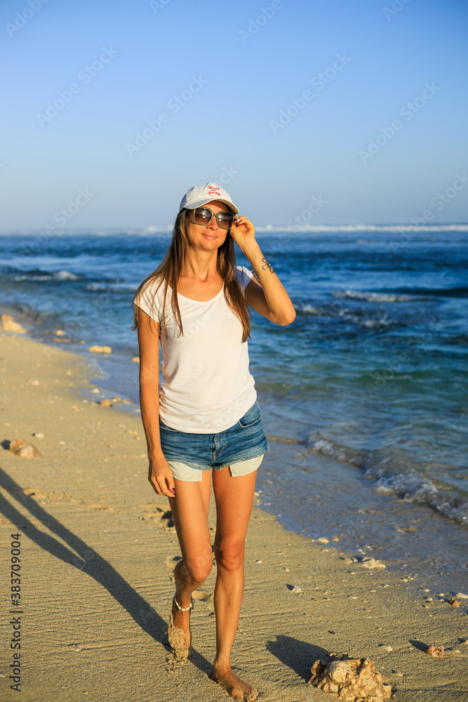 Slim young woman walking barefoot along the beach. Caucasian woman wearing jeans shorts, white T-shirt, cap, sunglasses. Happiness and freedom. Travel lifestyle. Bali, Indonesia