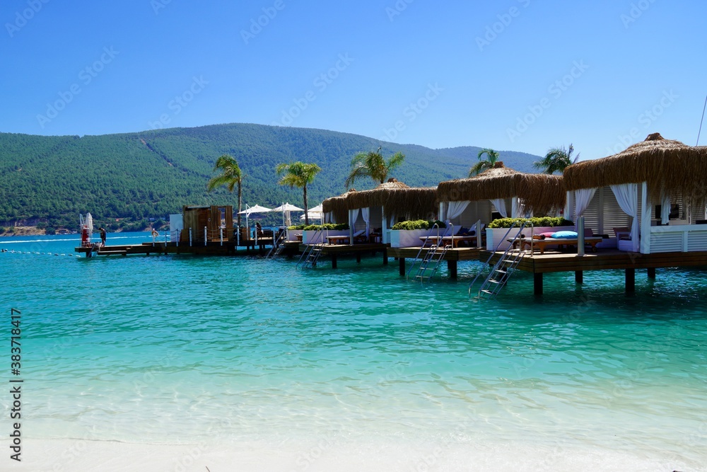 Bodrum, Turkey - August, 2020: Best beaches of Aegean Sea - tranquil organized beach. Bungallow on the water. Islands of Turkey.Pure emerald water. Bamboo houses bungalows for rest on the water