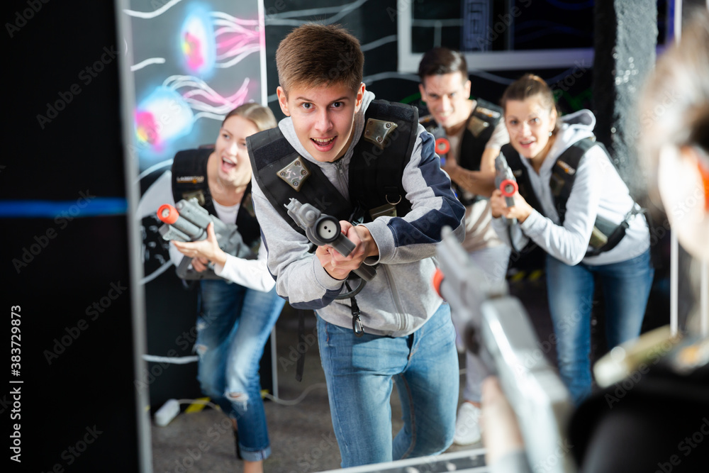 Cheerful guy aiming laser gun at other players during lasertag game in dark room..