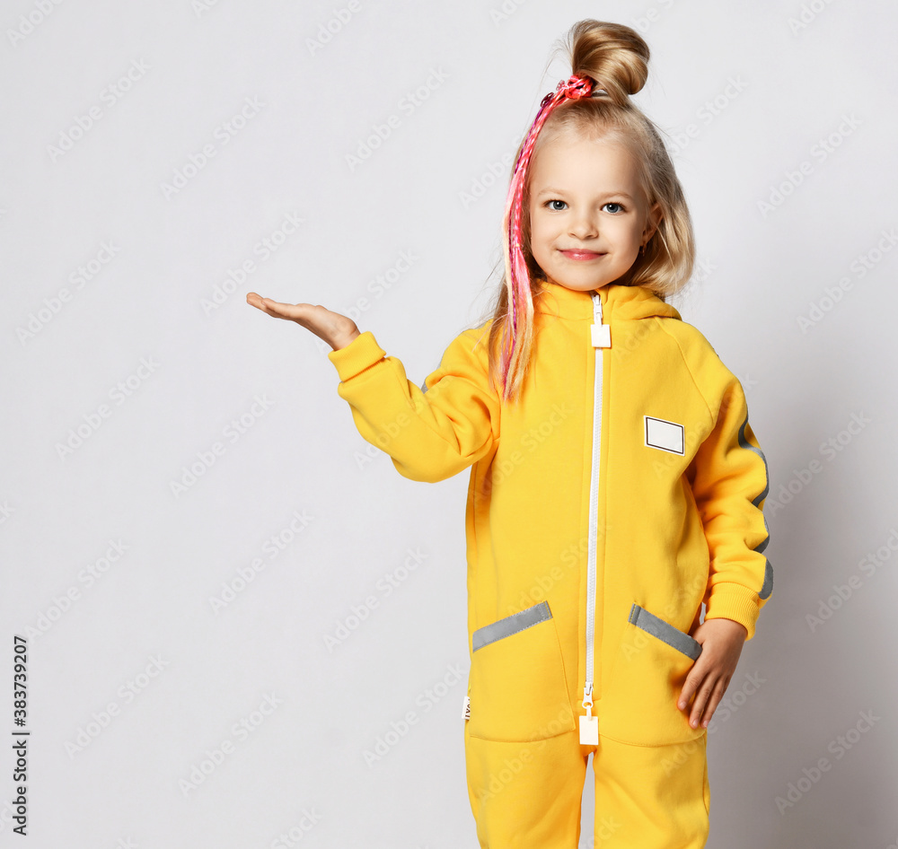 Smiling blonde kid baby girl with modern hairstyle and braids in yellow jumpsuit stands and holds hand up with open palm Smiling blonde kid baby girl with modern hairstyle and braids in yellow jumpsuit stands and holds hand up with open palm