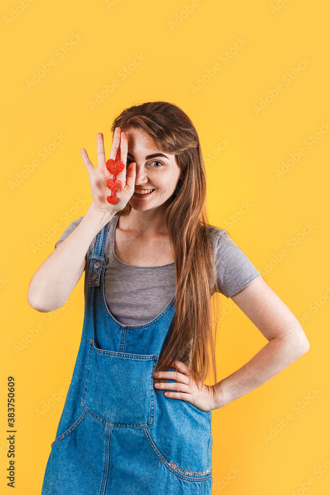 caucasian blonde girl in denim dress looking straight. close up. covers her face with red hearts in her hand. isolated on the yellow background. caucasian blonde girl in denim dress looking straight. close up. covers her face with red hearts in her hand. isolated on the yellow background.