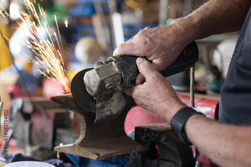 A man wearing a face mask protetion using an angle grinder in a garage workshop to do metal work fix create repair industrial job employment small local business sparks fire flames metal build constru photo