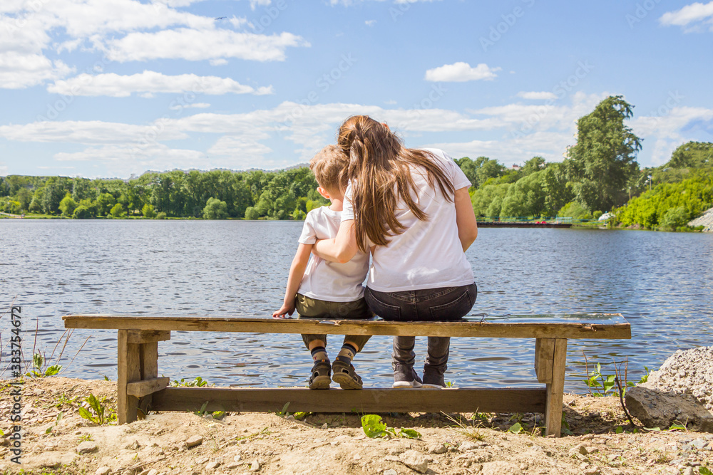 Mom and son cuddling sitting on bench by river shore at sunny summer day. Back view. Lifestyle, mothers tenderness. Son's day or mother's day concept