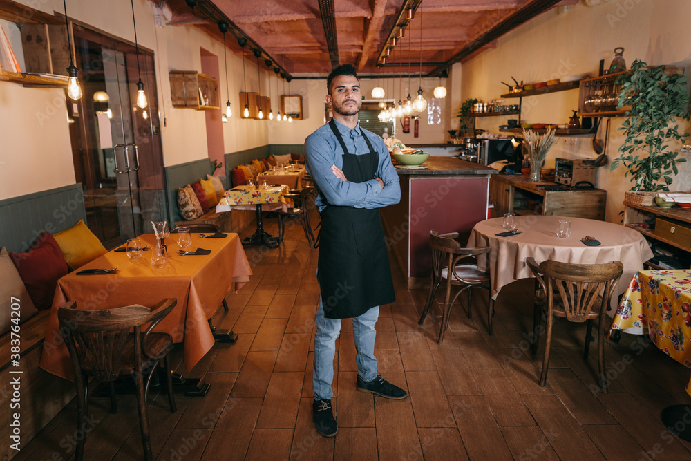 Serious confident handsome bearded business man crossing arms on chest and standing in the dining room of a modern restaurant. African american waiter looking at camera with determination. Serious confident handsome bearded business man crossing arms on chest and standing in the dining room of a modern restaurant. African american waiter looking at camera with determination.