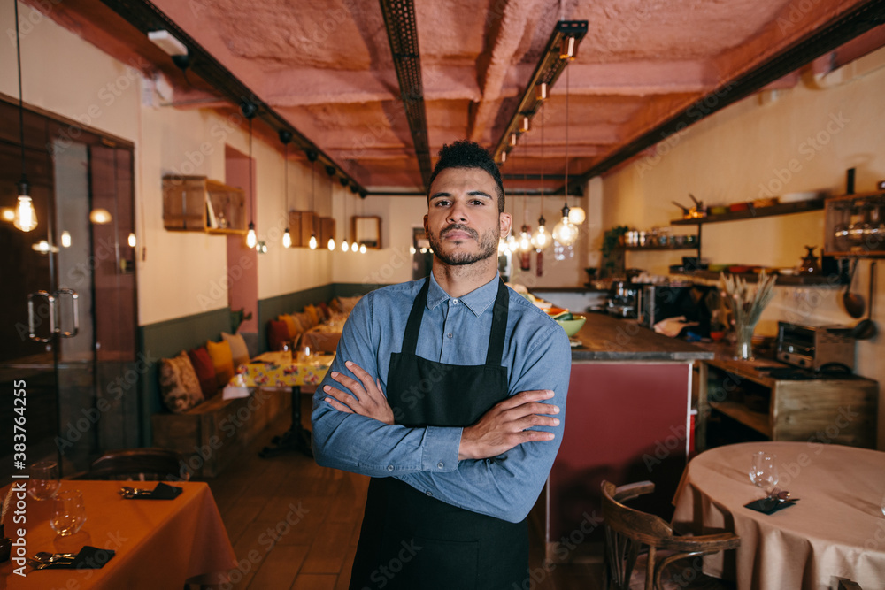 Fototapeta premium Portrait of young african american businessman standing in the dining room of his restaurant. Proud restaurant owner with his arms crossed and looking at camera. Small business concept