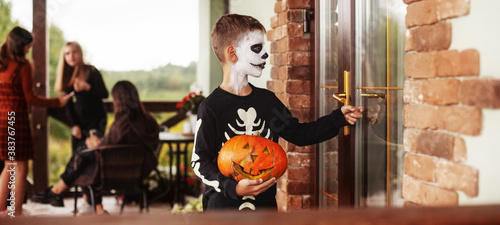 Boy and children in costumes and holiday make-up with pumpkin lantern Jack knocking on the door for sweets at halloween party outdoors near the house