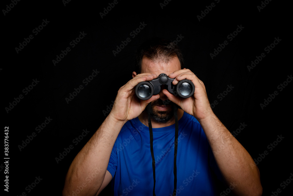 Bearded man dressed in blue t-shirt with binoculars posing against black background Bearded man dressed in blue t-shirt with binoculars posing against black background