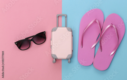Travel or beach resort flat lay. Mini plastic travel suitcase and flip flops, sunglasses on pink blue pastel background. Minimal style. Top view