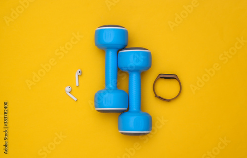 Dumbbells and gadgets for sports (smart bracelet and wireless headphones) on yellow background. Top view