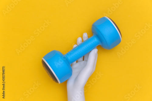 White mannequin hand holds blue plastic dumbbells on yellow background. Sport and fitness concept