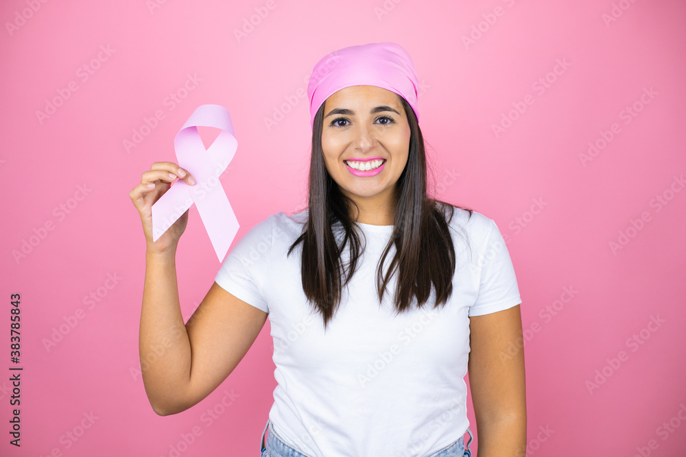 Young beautiful woman wearing pink headscarf holding brest cancer ribbon over isolated pink background with a happy face standing and smiling with a confident smile showing teeth Young beautiful woman wearing pink headscarf holding brest cancer ribbon over isolated pink background with a happy face standing and smiling with a confident smile showing teeth