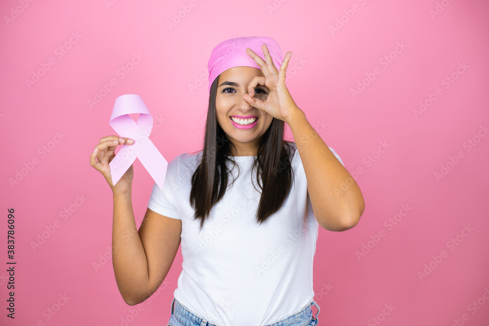 Young beautiful woman wearing pink headscarf holding brest cancer ribbon over isolated pink background doing ok gesture with hand smiling, eye looking through fingers with happy face. Young beautiful woman wearing pink headscarf holding brest cancer ribbon over isolated pink background doing ok gesture with hand smiling, eye looking through fingers with happy face.