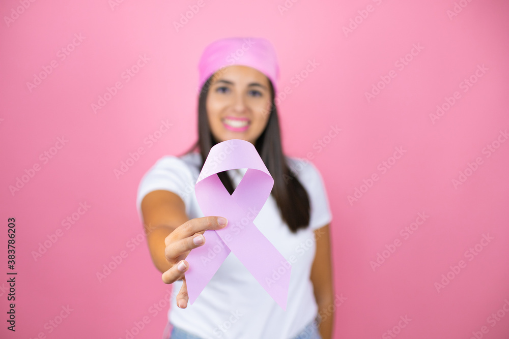 Young beautiful woman wearing pink headscarf holding brest cancer ribbon over isolated pink background with a happy face standing and smiling with a confident smile showing teeth Young beautiful woman wearing pink headscarf holding brest cancer ribbon over isolated pink background with a happy face standing and smiling with a confident smile showing teeth