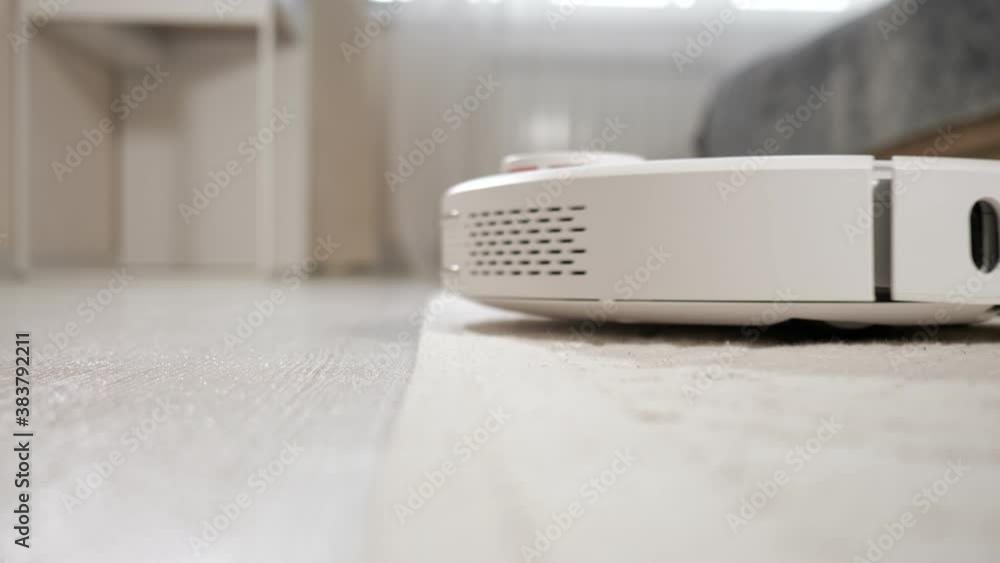 White automatic robot vacuum cleaner with small brushes hoovers wooden floor driving onto carpet in brightly lit room extreme closeup White automatic robot vacuum cleaner with small brushes hoovers wooden floor driving onto carpet in brightly lit room extreme closeup