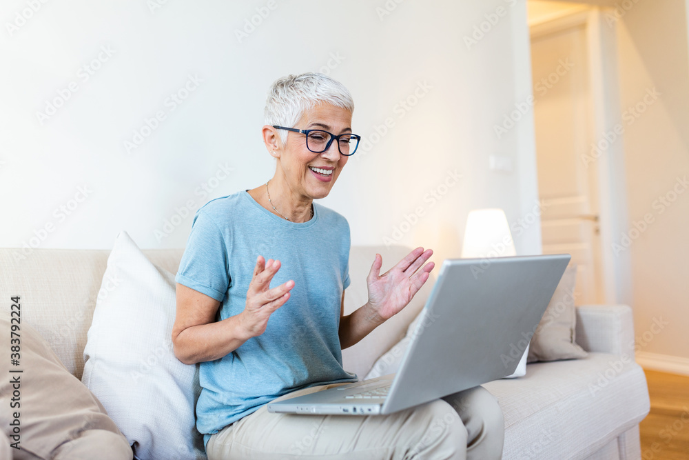 Happy mature woman waving to someone while having a video call over laptop at home. Gray-haired senior woman waving hand in front of laptop while having video call with her family members. Happy mature woman waving to someone while having a video call over laptop at home. Gray-haired senior woman waving hand in front of laptop while having video call with her family members.