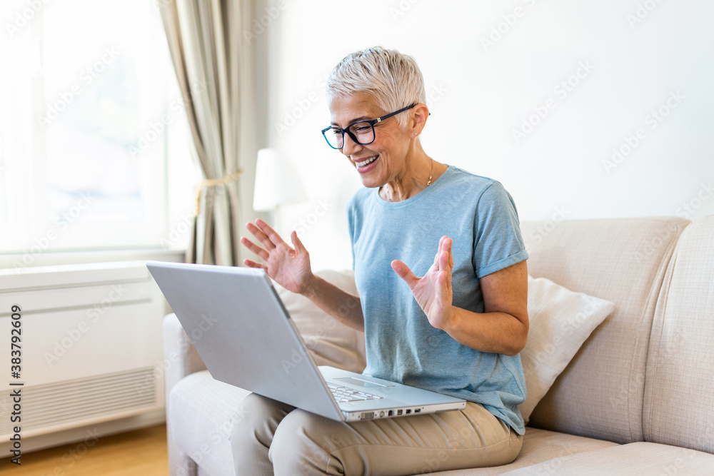 Happy mature woman waving to someone while having a video call over laptop at home. Gray-haired senior woman waving hand in front of laptop while having video call with her family members. Happy mature woman waving to someone while having a video call over laptop at home. Gray-haired senior woman waving hand in front of laptop while having video call with her family members.