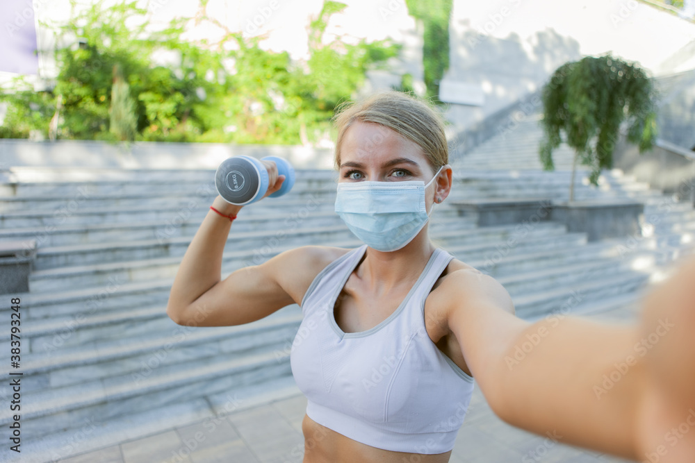 Selfie portrait of sportwoman in sportswear and medical protective face mask with dumbbells outdoors. Fitness classes during covid-19