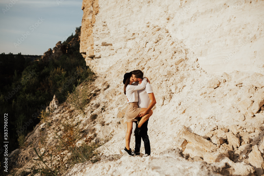 Bearded man and stylish woman in hat hug and kiss each other while travelling in mountain. Bearded man and stylish woman in hat hug and kiss each other while travelling in mountain.