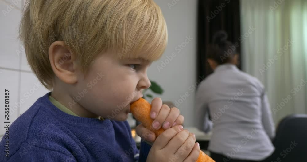 Cute Little Child Eats Big Carrot Sitting on Kitchen Table while Mother Cooks Food at Home 4K Cute Little Child Eats Big Carrot Sitting on Kitchen Table while Mother Cooks Food at Home 4K