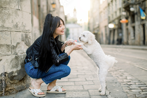 Young Asian urban girl wearing stylish casual clothes, jeans and leather jacket, walks with little cute white dog on the street against the background of the wall, and old vintage buildings