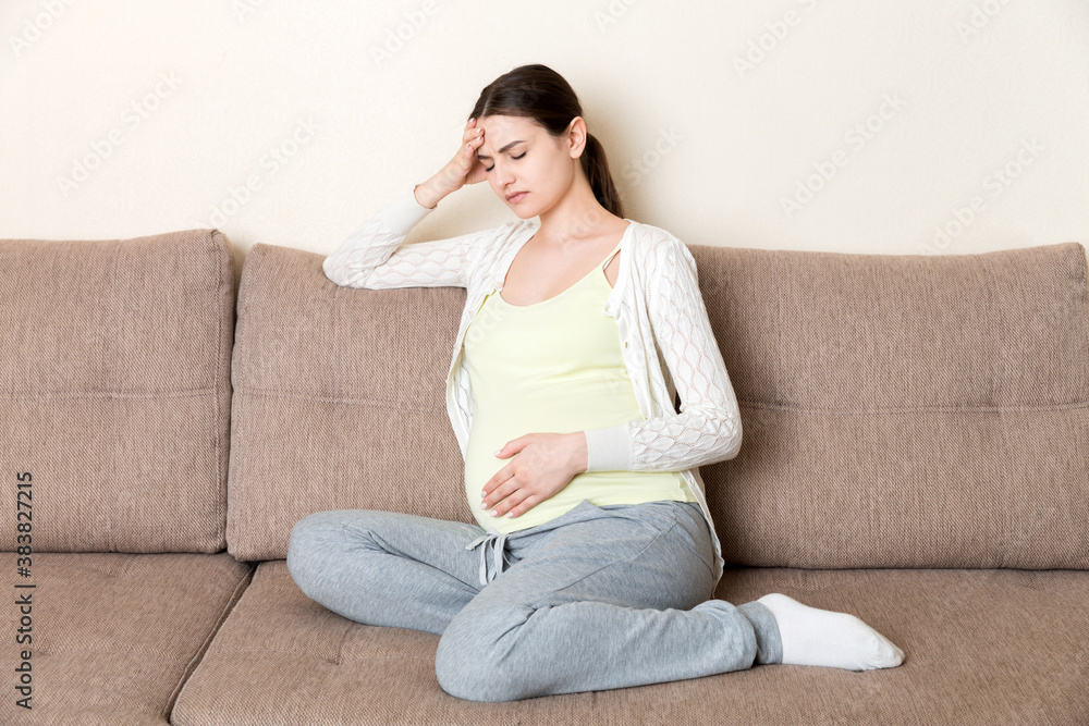 Young pregnant woman with headache sitting on sofa. Pregnancy expectation concept Young pregnant woman with headache sitting on sofa. Pregnancy expectation concept