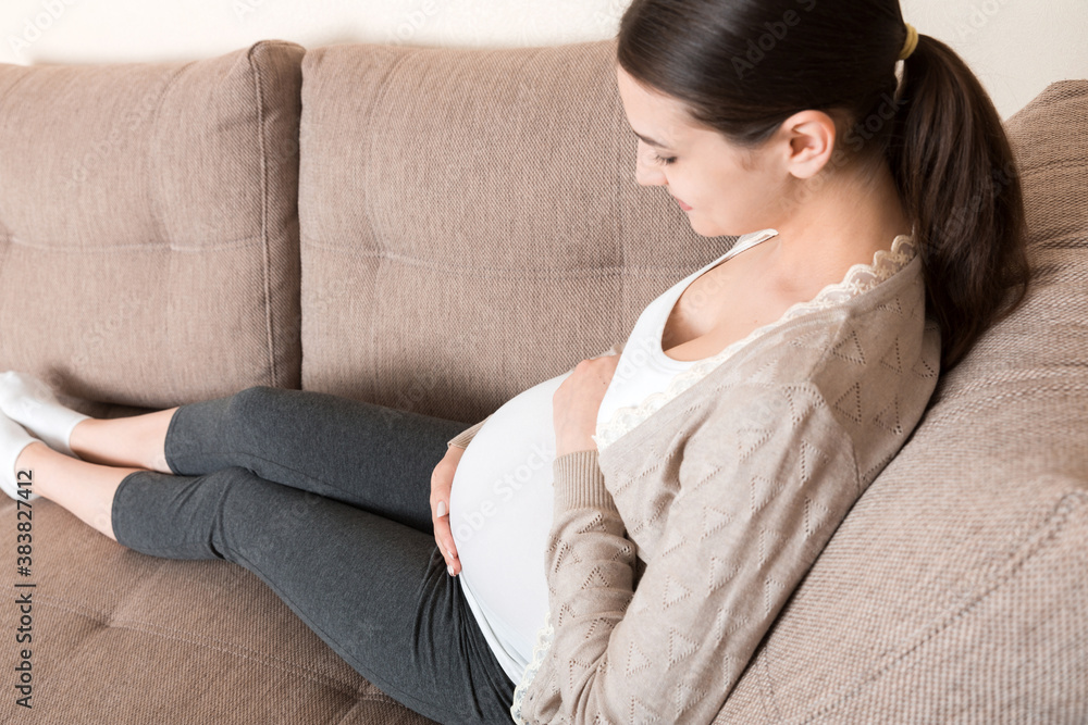 photo of cute caucasian future mom lying in light living room on sofa and holding hands on tummy photo of cute caucasian future mom lying in light living room on sofa and holding hands on tummy