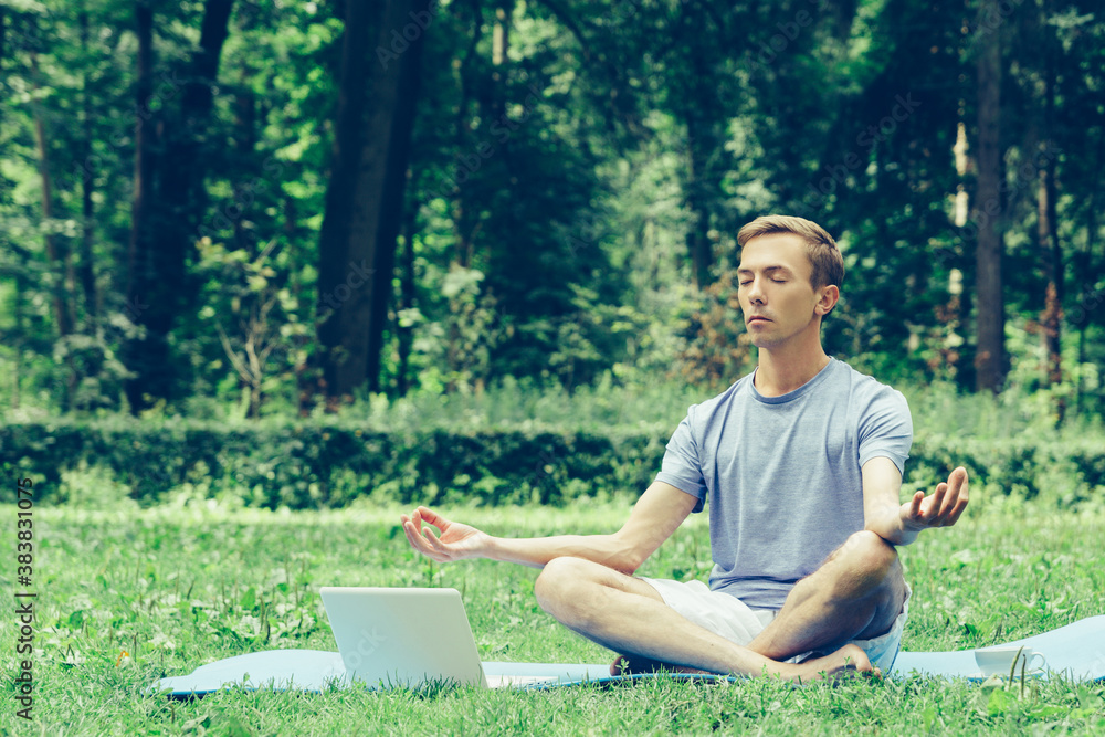 Obraz premium Young attractive man in sport clothes is meditating in the lotus position with a pacified face in the park against the background of green grass and trees.
