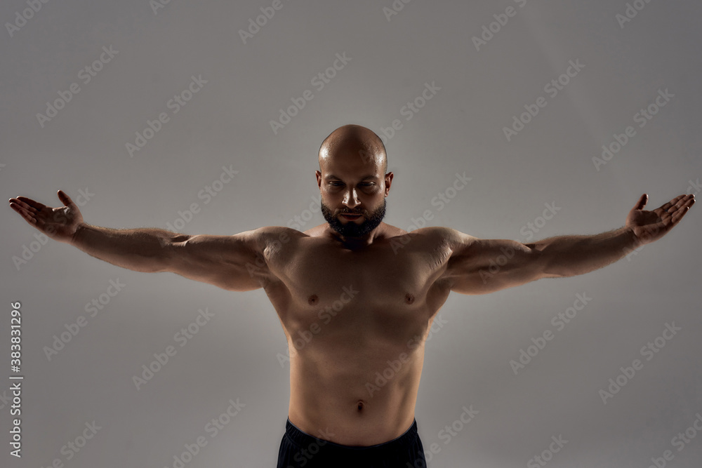 Professional bodybuilder posing. Muscular strong bald man with naked torso standing with outstretched arms isolated over grey background Professional bodybuilder posing. Muscular strong bald man with naked torso standing with outstretched arms isolated over grey background