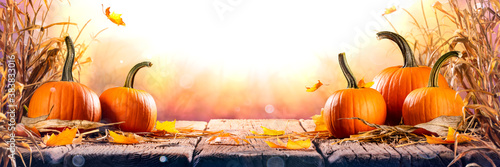 Εκτύπωση καμβά Pumpkins With Cornstalks And Falling Leaves On Wooden Harvest Table At Sunset -