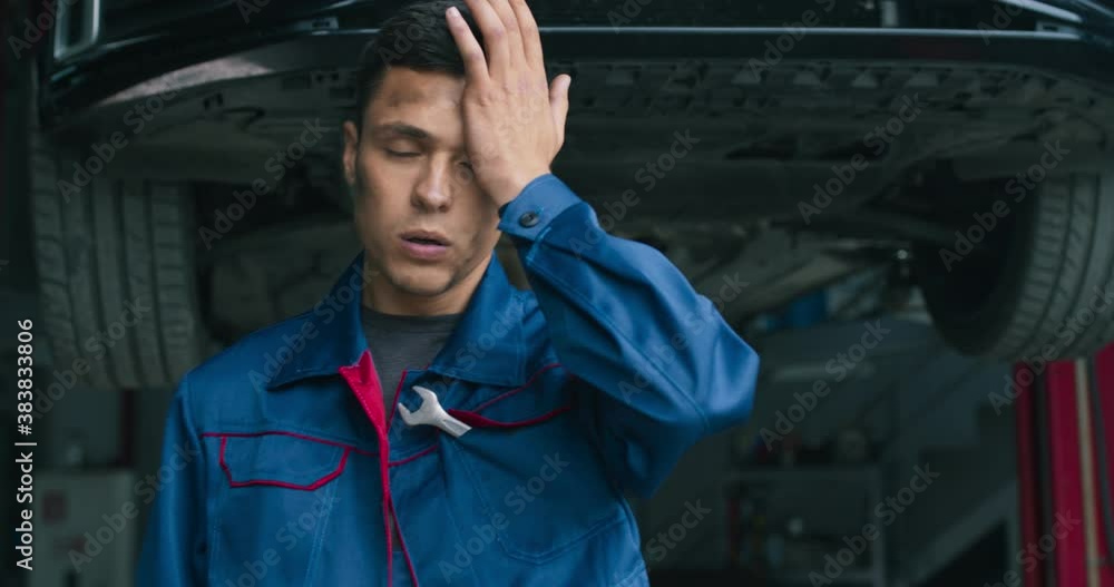 Young mechanic tired after he finished to fix car, rubbing his forehead, coming out of lifted vehicle Young mechanic tired after he finished to fix car, rubbing his forehead, coming out of lifted vehicle