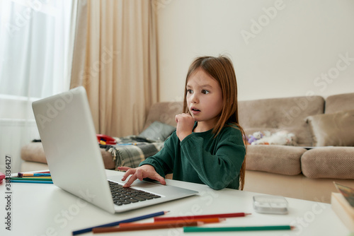 A cute little girl expressing an astonishment by looking at a laptop's screen sitting alone at a table during distance education