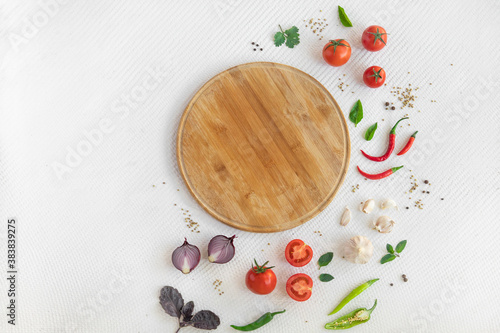 Empty round wooden cutting board on white textured background with spices, onion, garlic, chilli pepper, cherry tomatoes, basilic and greens with copy space. Top view.
