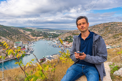 Fotografie Handsome middle aged tourist man sitting on stone against beautiful landscape of small coastal town in cloudy day