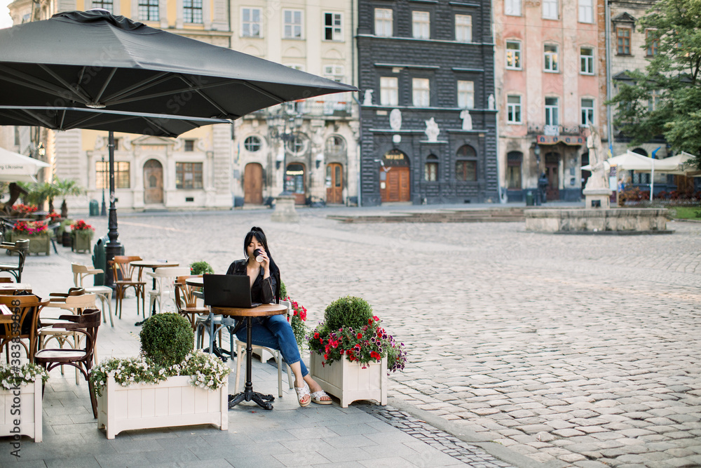 Obraz premium Young attractive brunette woman using laptop at outdoor city cafe, sitting at the table and drinking coffee. Old vintage buildings of European city on the background