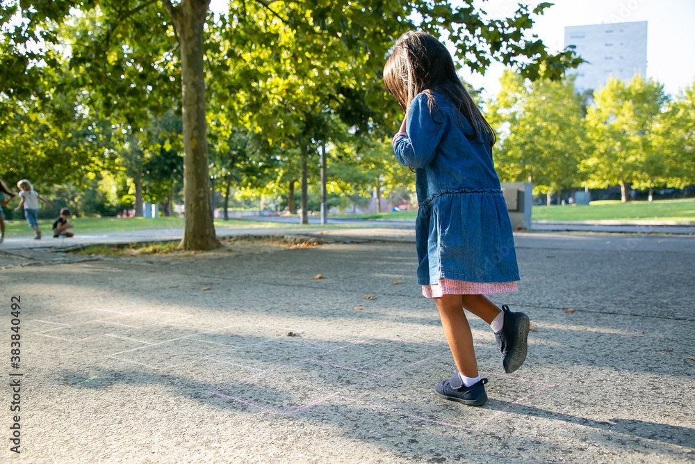 Adorable black haired little girl playing hopscotch in city park. Full length, copy space. Childhood concept Adorable black haired little girl playing hopscotch in city park. Full length, copy space. Childhood concept