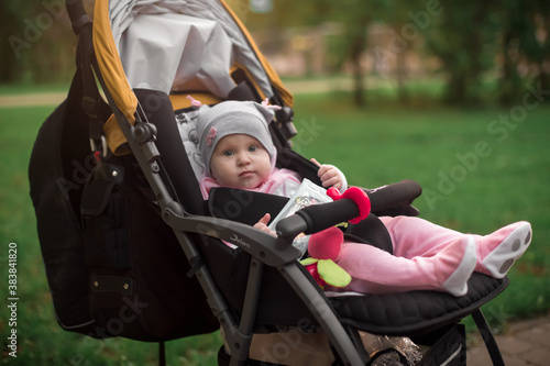 A girl in pink clothes is sitting in a yellow stroller in a Park among green trees.