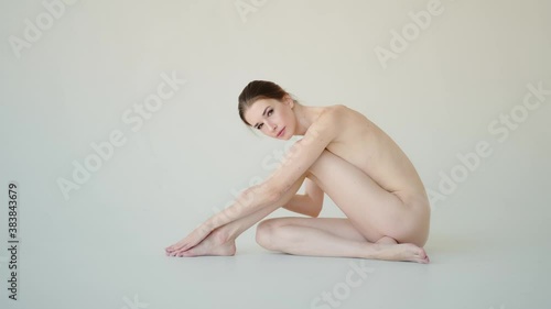 Young nude slim woman is sitting and touching legs. Portrait of a naked skinny woman on a white background photo