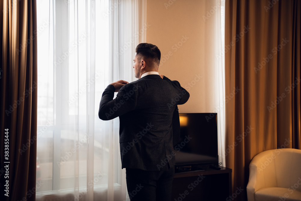 Serious young businessman standing near the window in his bedfoom, fixing his jacket, getting ready to go out. Serious young businessman standing near the window in his bedfoom, fixing his jacket, getting ready to go out.