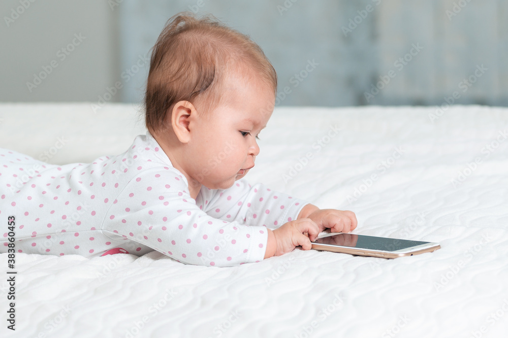 Portrait of baby is lying on the bed and playing with a smartphone. Side view. Copy space. Concept of children's games with modern devices Portrait of baby is lying on the bed and playing with a smartphone. Side view. Copy space. Concept of children's games with modern devices