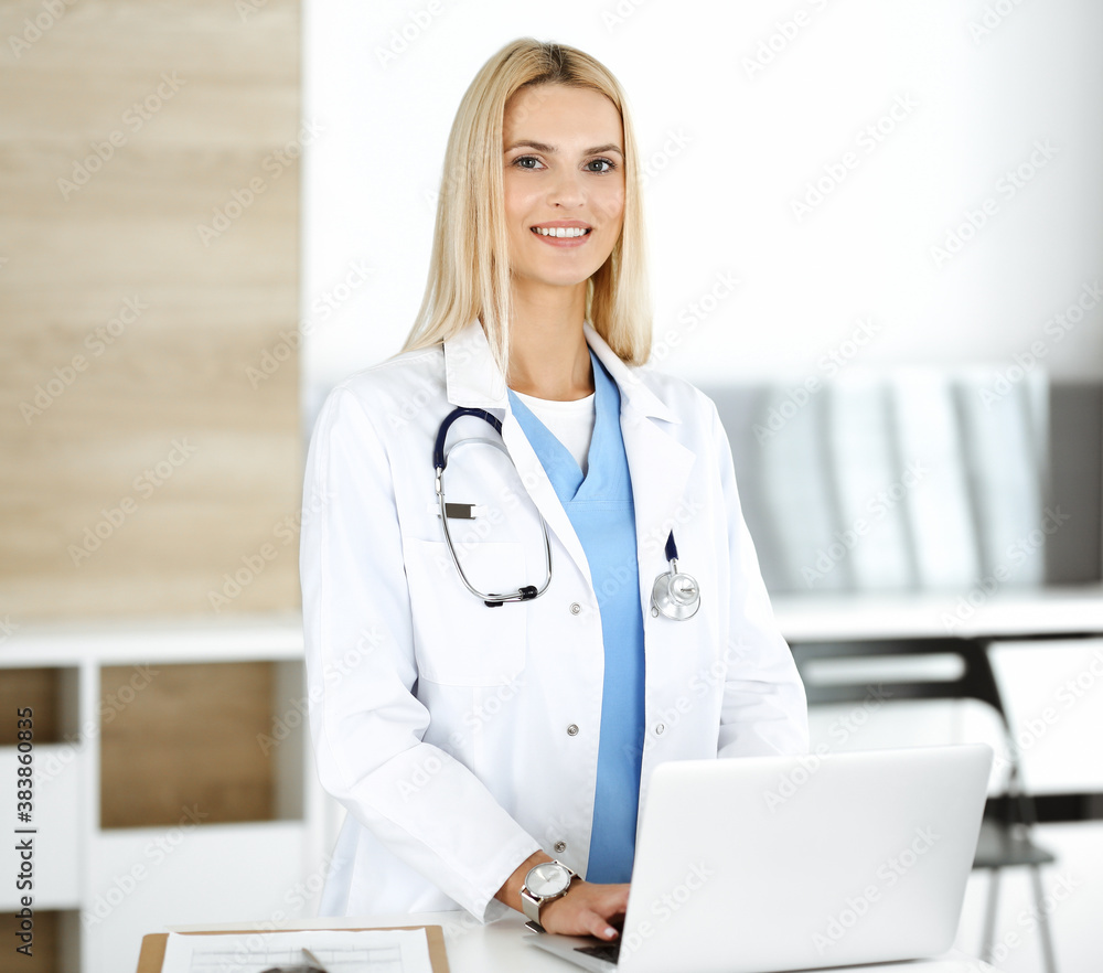 Blond female physician is lsmiling while using laptop computer. Woman-doctor at work in clinic excited and happy of her profession. Medicine concept Blond female physician is lsmiling while using laptop computer. Woman-doctor at work in clinic excited and happy of her profession. Medicine concept