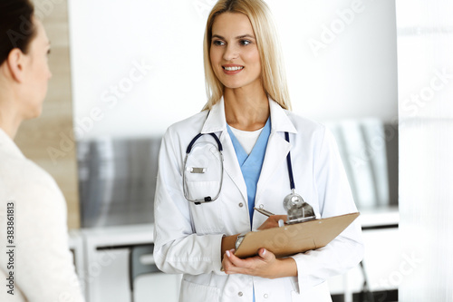 Woman-doctor at work in hospital is happy to consult female patient. Blonde physician checks medical history record and exam results while using clipboard. Medicine concept