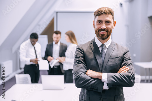 Portrait of a positive looking young business professional standing with his arms crossed with colleagues talking in the background.