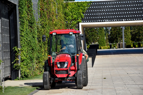 A red tractor stands in the park. A tractor is standing on the laid paving slabs. Tractor with driver at work in the park.