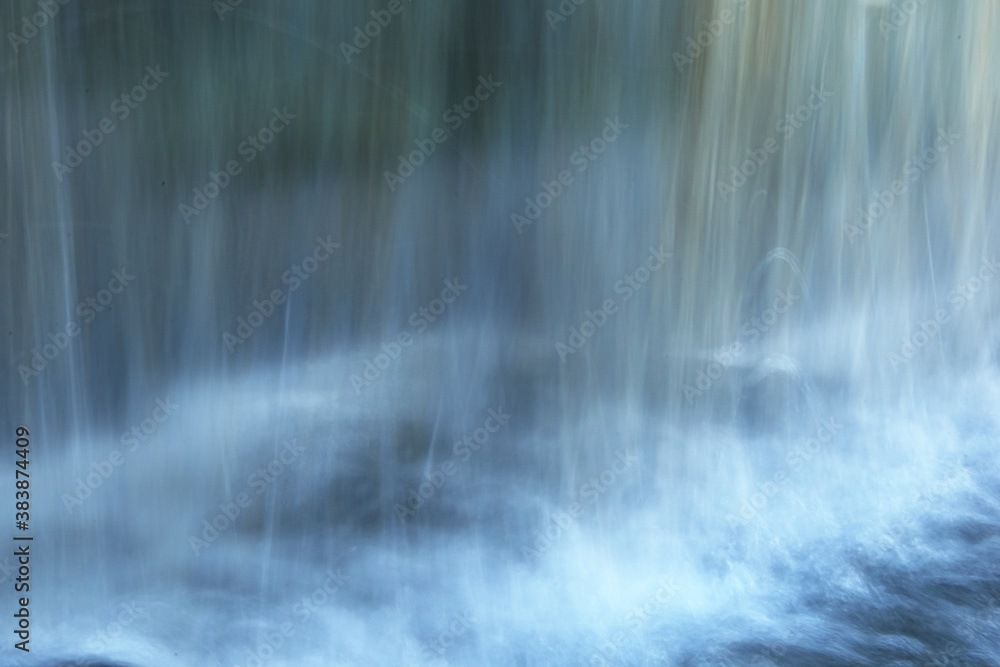 Water flows down a small wall with reflections behind. Blue-white blurred water background. Blurred artificial waterfall background. Water flows down a small wall with reflections behind. Blue-white blurred water background. Blurred artificial waterfall background.